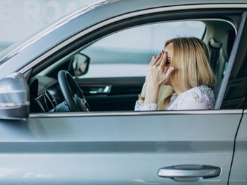 Eine gestresste Frau sitzt auf dem Fahrersitz eines Autos und bedeckt ihr Gesicht mit den Händen, was Frustration oder Angst widerspiegelt.