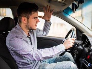 Eine Person fährt ein Auto, hat eine Hand am Lenkrad und hebt die andere in einer Geste der Frustration.